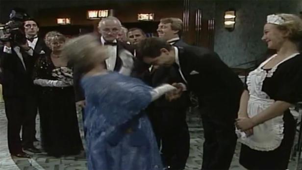 Eine Frau im blauen Kleid schlägt einen Mann im Smoking, während andere zusehen.