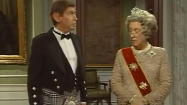 Ein Mann im Kilt und eine Frau mit Krone und Schärpe stehen in einem Raum.