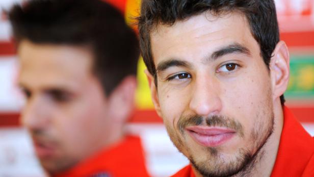 Ein Mann mit Bart lächelt in die Kamera, eine weitere Person ist im Hintergrund unscharf zu sehen.