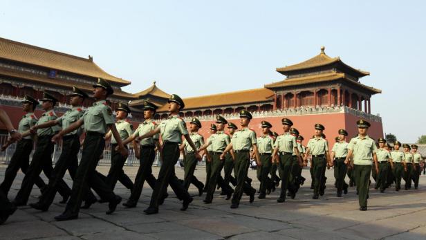 Eine Reihe chinesischer Soldaten marschiert auf einem Platz vor traditionellen chinesischen Gebäuden.