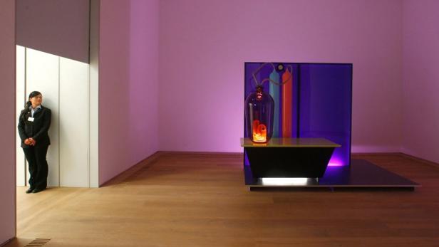 Eine Frau steht in einem Ausstellungsraum neben einer Installation mit Glasbehälter und farbigen Röhren.