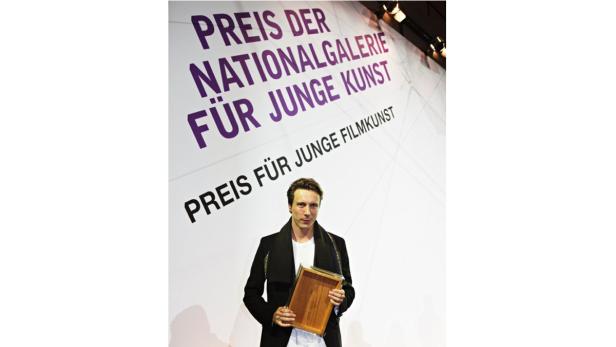 Ein Mann hält eine Auszeichnung vor dem Hintergrund des „Preis der Nationalgalerie für junge Kunst“.