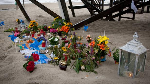 Ein improvisiertes Denkmal am Strand mit Blumen, Windrädern und Kerzen.