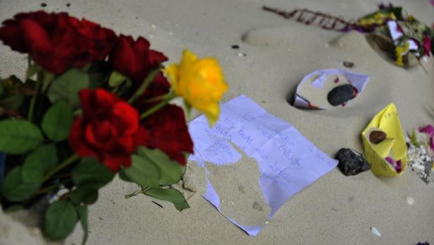 Blumen, ein Zettel mit einer handschriftlichen Nachricht und Papierboote liegen im Sand.