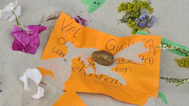 Eine orangefarbene Notiz mit der Aufschrift „Viel Glück“ und dem Namen Sebastian liegt im Sand.