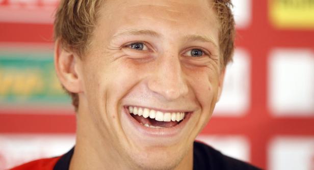 Ein lachender Mann mit blonden Haaren und blauen Augen vor einem roten Hintergrund.