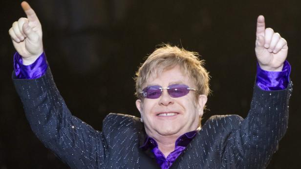 Elton John mit erhobenen Armen und lila Sonnenbrille auf der Bühne.