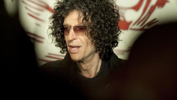 Howard Stern mit lockigem Haar und Sonnenbrille vor einem gemusterten Hintergrund.
