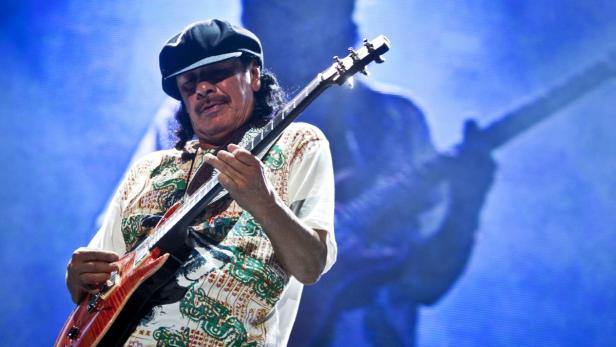 Carlos Santana spielt Gitarre auf einer Bühne mit blauem Hintergrund.