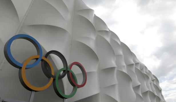 Die olympischen Ringe vor einer weißen Fassade mit Wolken im Hintergrund.