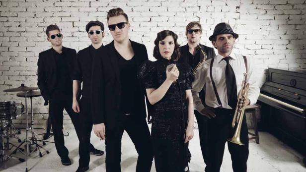 Eine Band mit Sängerin posiert vor einer weißen Ziegelwand mit Musikinstrumenten.