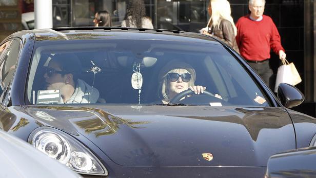 Eine Frau mit Hut und Sonnenbrille fährt einen dunklen Porsche, während ein Mann auf dem Beifahrersitz sitzt.