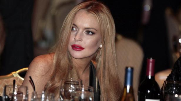Lindsay Lohan sitzt an einem Tisch mit Weingläsern und Weinflaschen.
