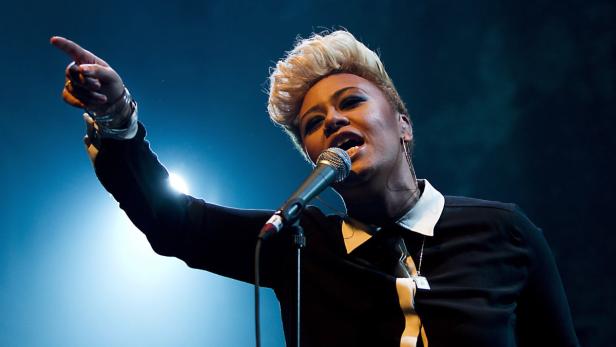 Eine Sängerin mit blonden Haaren singt in ein Mikrofon und zeigt mit dem Finger nach oben.