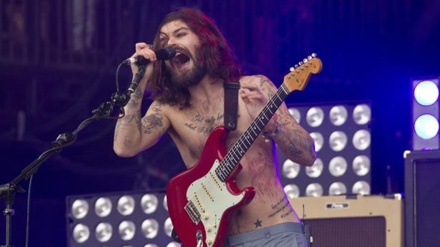 Ein Mann mit Tätowierungen singt und spielt Gitarre auf einer Bühne.