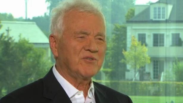 Ein älterer Mann mit weißem Haar und dunklem Anzug vor einem Fenster mit Blick auf ein Haus.