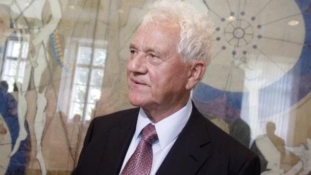 Ein Mann mit weißem Haar, Anzug und Krawatte vor einem gemusterten Hintergrund.