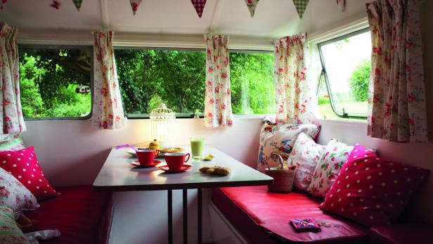 Innenraum eines Wohnwagens mit Tisch, Geschirr und floralen Vorhängen.