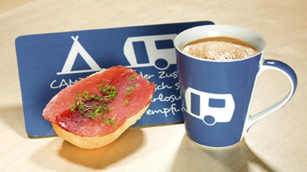 Ein Brötchen mit Schinken und Kräutern neben einer Tasse Kaffee auf einem Camping-Untersetzer.