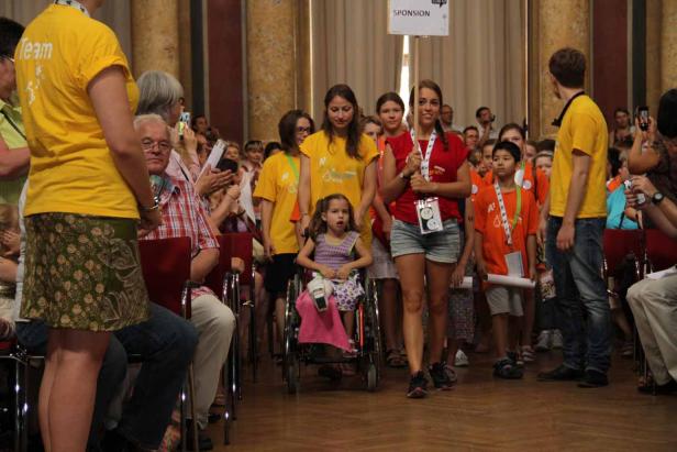 Eine junge Frau schiebt ein Mädchen im Rollstuhl, gefolgt von einer Gruppe von Menschen.