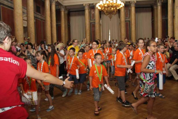 Eine Gruppe Kinder in orangefarbenen T-Shirts geht durch einen großen Saal mit Säulen und einem Kronleuchter.