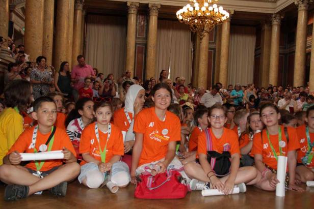 Eine Gruppe von Kindern sitzt in einem großen Saal mit Säulen und einem Kronleuchter.
