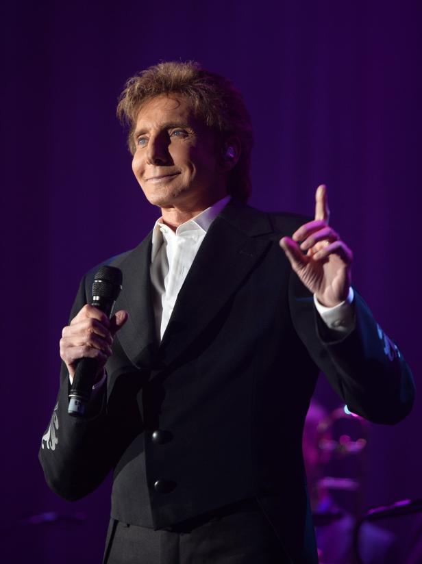 Barry Manilow hält ein Mikrofon und zeigt mit dem Finger nach oben.