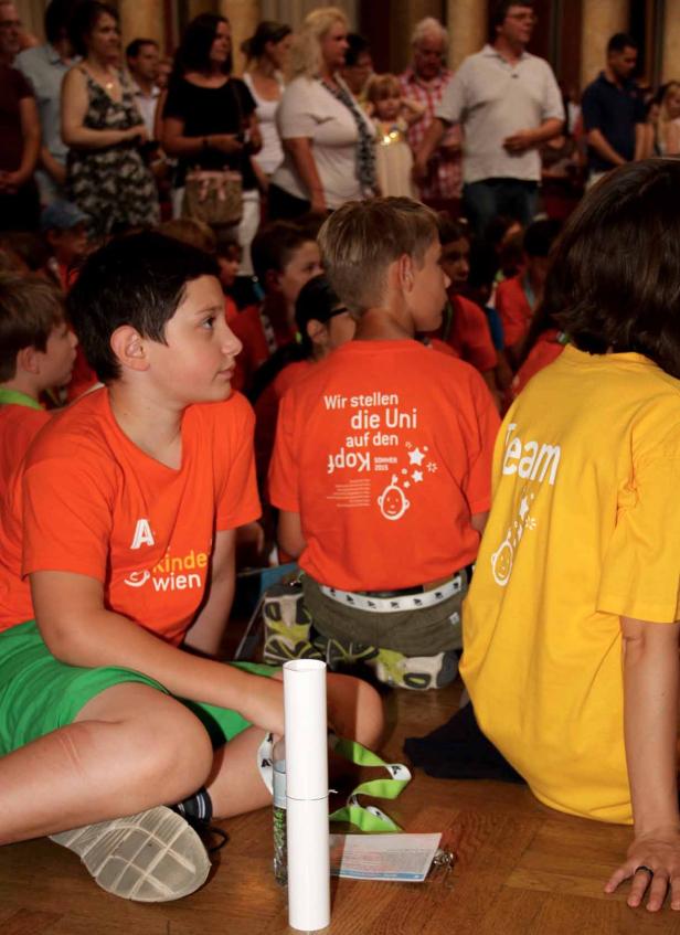 Kinder sitzen auf dem Boden, einige tragen T-Shirts mit der Aufschrift „Wir stellen die Uni auf den Kopf“.