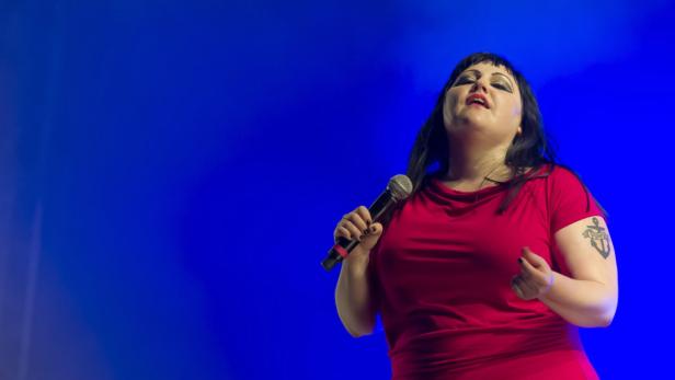 Beth Ditto singt in einem roten Kleid vor einem blauen Hintergrund.