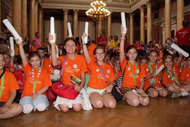 Eine Gruppe Kinder in orangefarbenen T-Shirts sitzt auf dem Boden und hält Papierrollen hoch.