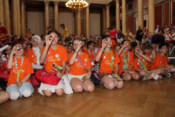 Eine Gruppe Kinder in orangefarbenen T-Shirts sitzt auf dem Boden und blickt durch kleine Linsen.