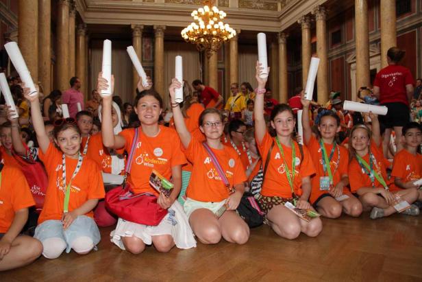 Eine Gruppe Kinder in orangefarbenen T-Shirts kniet und hält Papierrollen hoch.