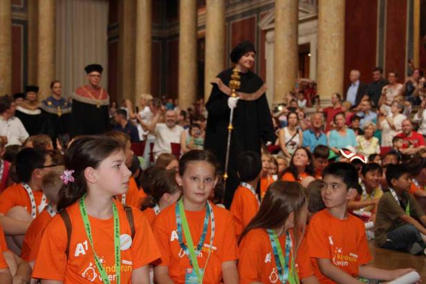 Eine Gruppe von Kindern sitzt vor einer Menschenmenge und Personen in akademischer Kleidung.