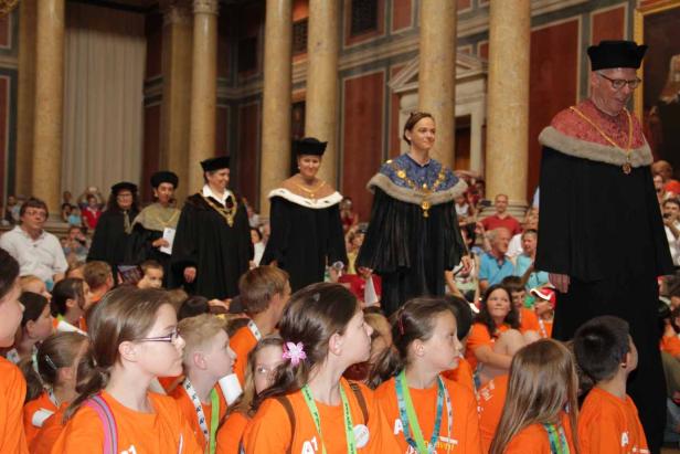 Eine Gruppe von Kindern beobachtet eine Prozession von Personen in akademischer Kleidung.