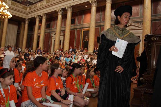 Eine Frau in akademischer Kleidung hält eine Rede vor einer Gruppe von Kindern in einem großen Saal.