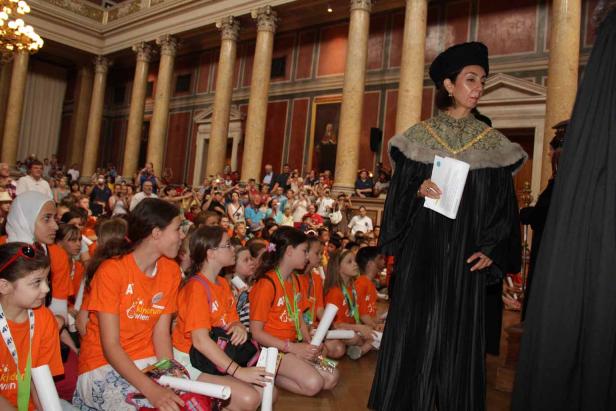 Eine Frau in akademischer Kleidung steht vor einer Gruppe von Kindern in orangefarbenen T-Shirts in einem großen Saal.