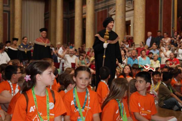 Eine Gruppe Kinder in orangefarbenen T-Shirts sitzt vor einer Menschenmenge und Würdenträgern in einer Halle.