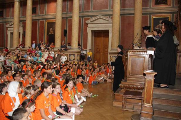 Eine Gruppe Kinder in orangefarbenen T-Shirts sitzt in einem Saal und hört einer Rede zu.