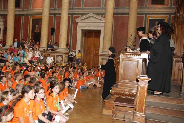 Eine Gruppe Kinder in orangefarbenen T-Shirts sitzt auf dem Boden und hört einer Rede zu.