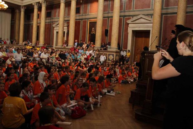 Eine große Gruppe von Kindern sitzt auf dem Boden in einem Saal mit Säulen.