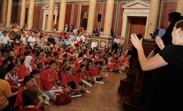 Eine große Gruppe von Kindern sitzt auf dem Boden in einem verzierten Saal und hört zu.