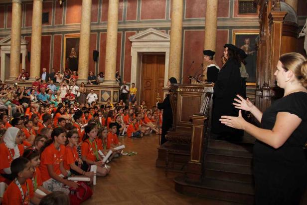 Eine Gruppe Kinder in orangefarbenen T-Shirts sitzt in einem großen Saal vor einer Bühne.