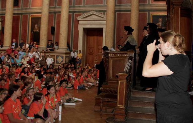 Eine Frau dolmetscht eine Rede vor einer Gruppe von Kindern in einem großen Saal.