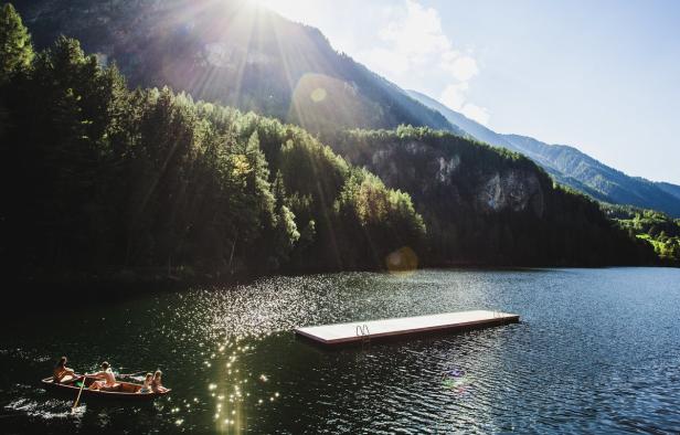 Eine Familie rudert auf einem Bergsee mit einem Steg im glitzernden Sonnenlicht.