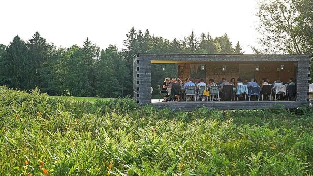Eine Gruppe von Menschen sitzt an einem langen Tisch in einem offenen Holzpavillon inmitten einer grünen Landschaft.