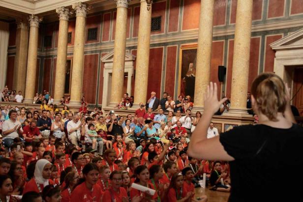 Eine Menschenmenge, darunter viele Kinder in roten T-Shirts, sitzt in einem großen Saal mit Säulen.