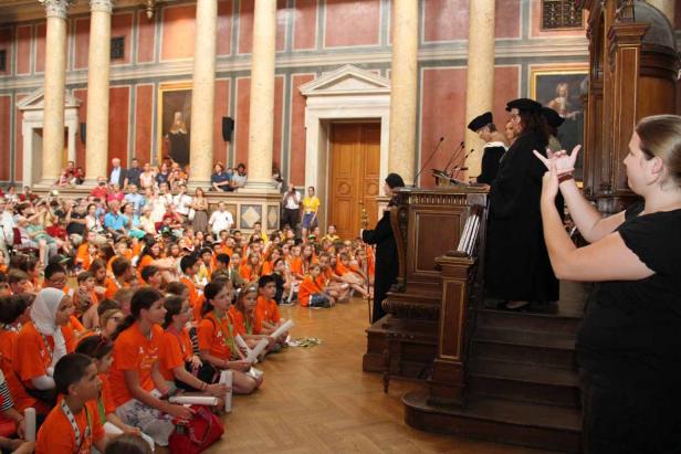 Eine Gruppe Kinder sitzt auf dem Boden, während Redner auf einem Podium sprechen und eine Frau in Gebärdensprache übersetzt.