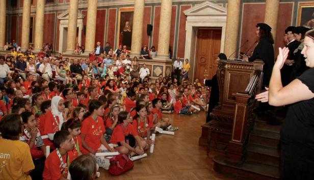 Eine Gruppe Kinder sitzt auf dem Boden und hört einer Rede in einem großen Saal zu.