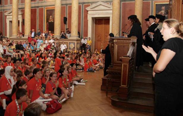 Eine Gruppe Kinder sitzt auf dem Boden und hört einer Rede in einem großen Saal zu.
