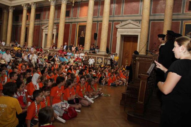 Eine Gruppe von Kindern sitzt auf dem Boden in einem großen Saal mit Säulen und hört einer Rede zu.